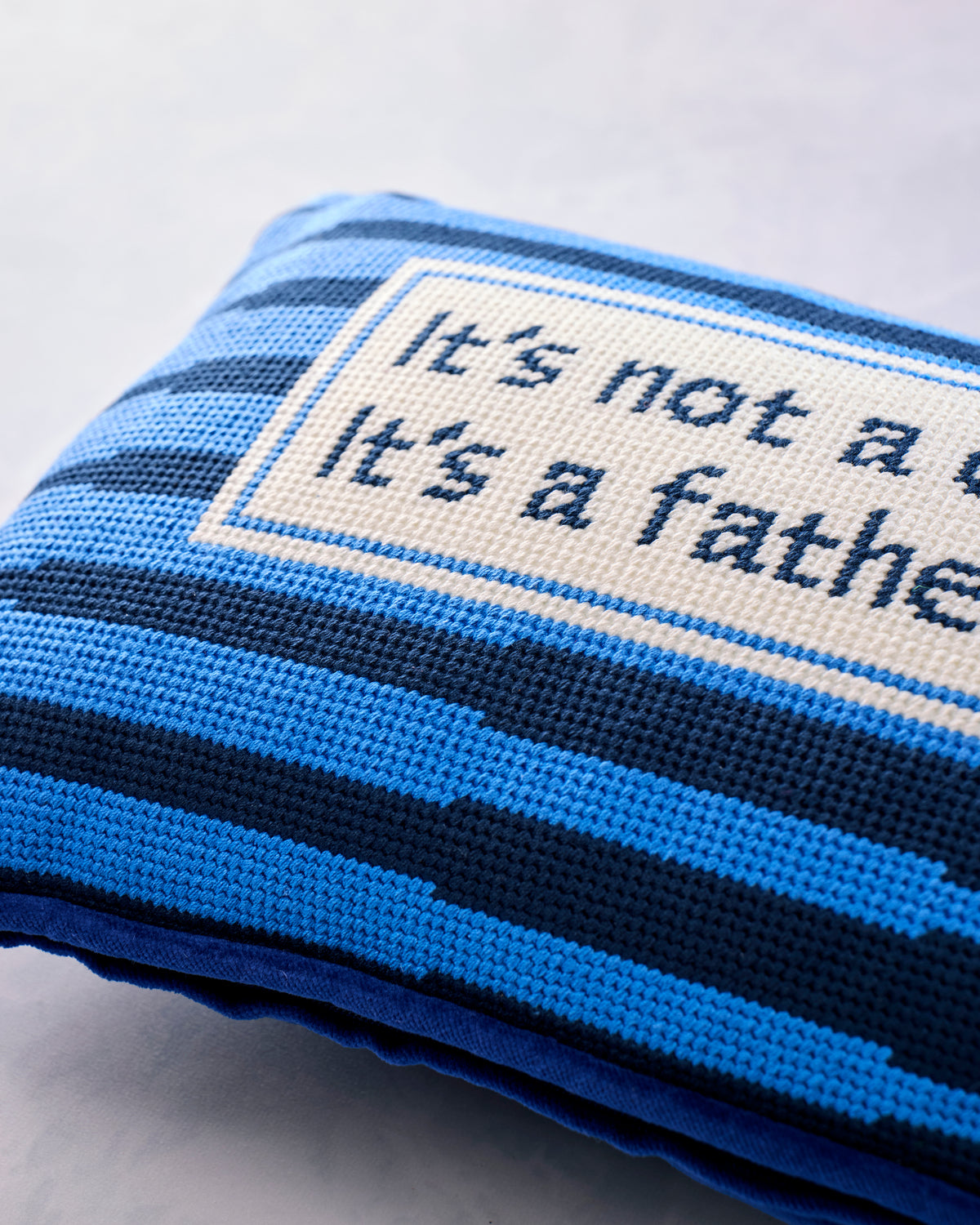 Close-up of blue and navy striped needlepoint stitching with a bordered text panel reading “It’s not a dad bod; it’s a father figure.”