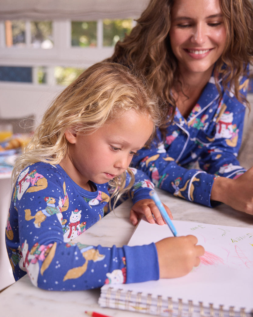 Child drawing at a table while an adult in matching blue winter-print pajamas looks on.
