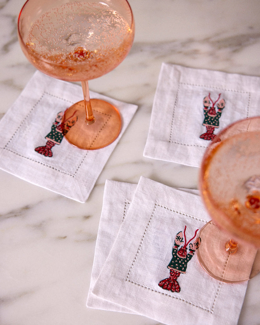 White embroidered cocktail napkins arranged on a marble surface with pink drinks in stemmed glasses.