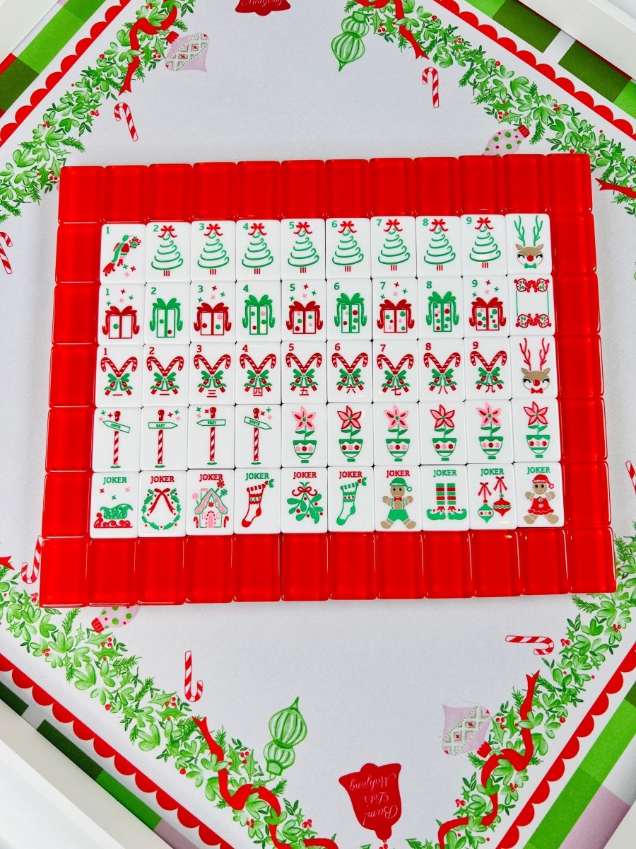 Holiday Mahjong tiles arranged in rows on red racks over the Holly Jolly Christmas mat, with Christmas tree, gift, candy cane, ornament, and holiday character designs.
