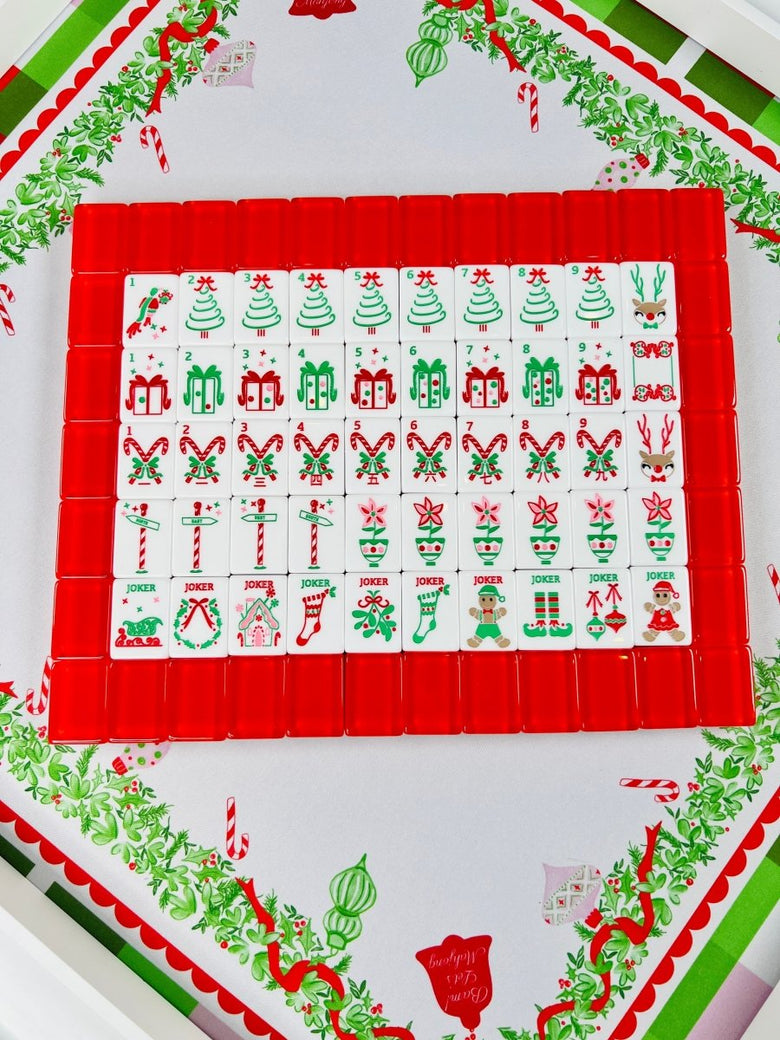 Holiday Mahjong tiles arranged in rows on red racks over the Holly Jolly Christmas mat, with Christmas tree, gift, candy cane, ornament, and holiday character designs.