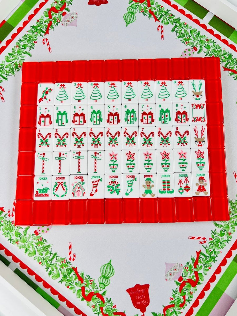 Holiday Mahjong tiles arranged in rows on red racks over the Holly Jolly Christmas mat, with Christmas tree, gift, candy cane, ornament, and holiday character designs.
