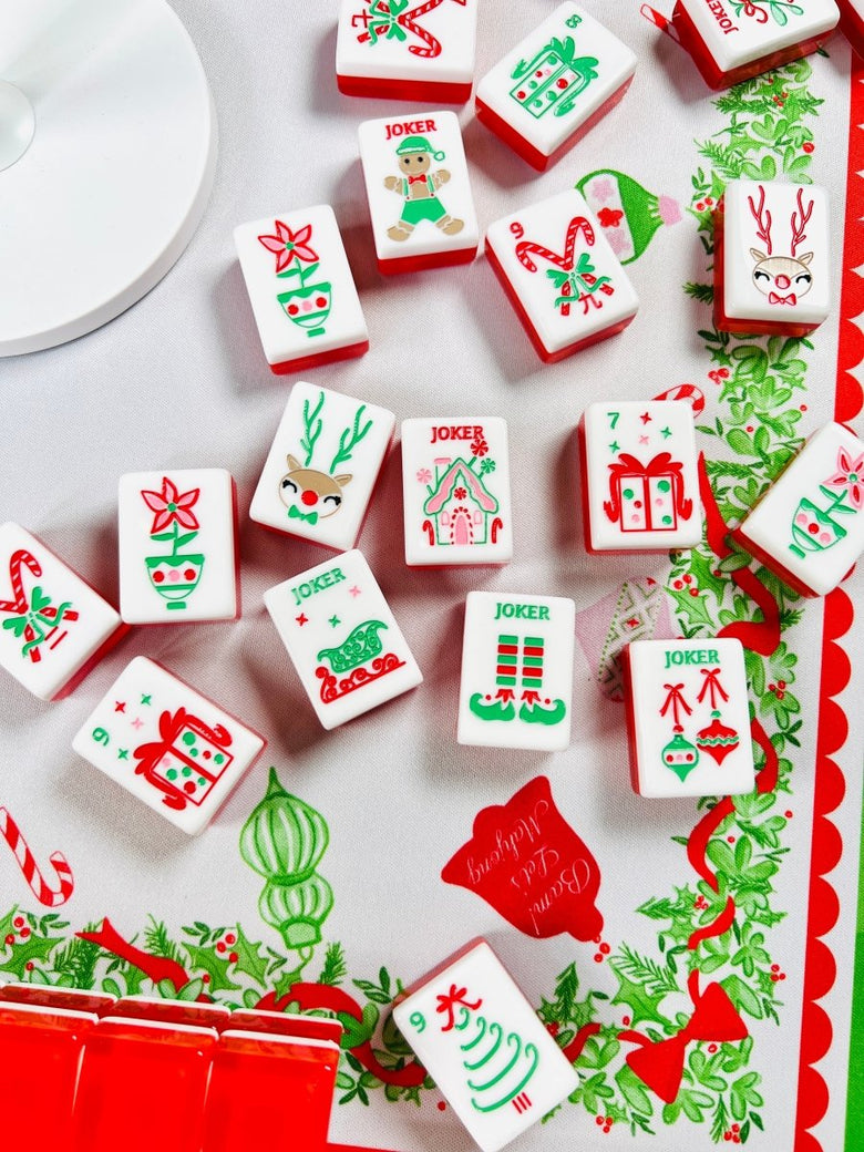 Close view of holiday mahjong tiles scattered on a Christmas table covering, including candy canes, wrapped gifts, poinsettias, ornaments, and joker tiles.