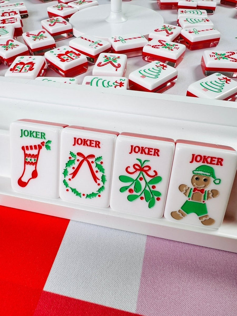 Holiday mahjong tiles scattered around a white circular stand, with upright joker tiles showing a stocking, wreath, mistletoe, and elf designs.