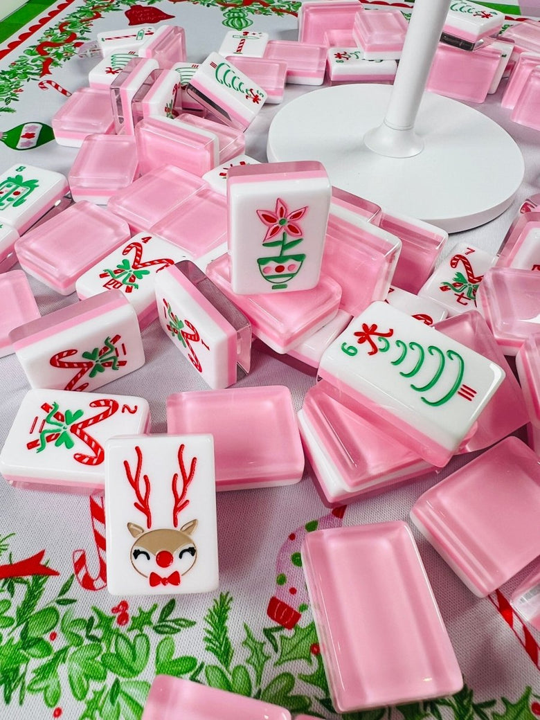 Holiday mahjong tiles displayed among open translucent pink cases, including upright poinsettia, candy cane, and reindeer designs.