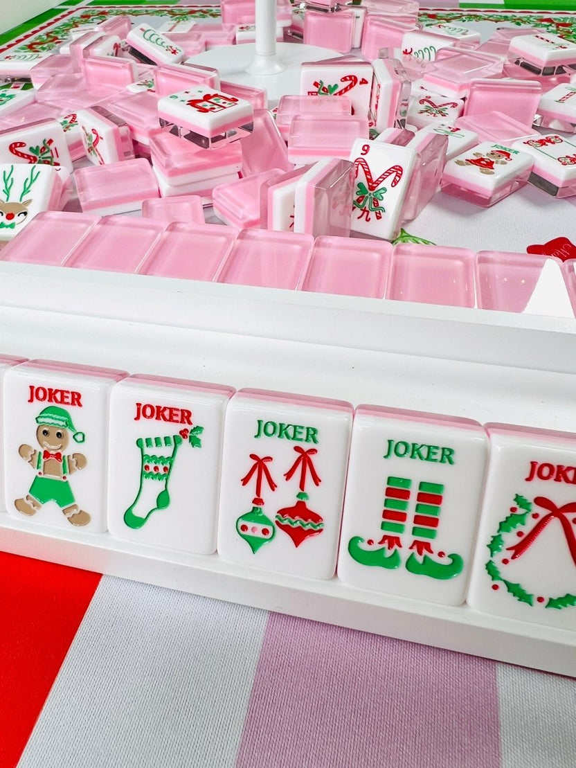Holiday mahjong tiles and open pink cases arranged around a white circular stand, with upright joker tiles showing an elf, stocking, ornaments, mustache, and wreath.