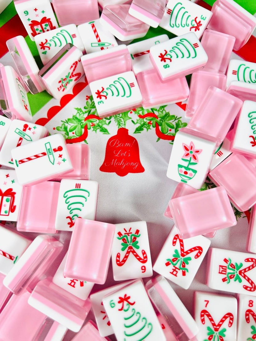 Holiday mahjong tiles and open translucent pink tile cases arranged on a Christmas table covering.