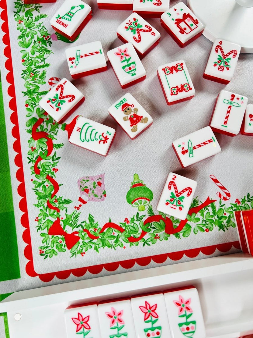 Holiday mahjong tiles scattered on a Christmas-themed table covering, showing candy canes, wrapped gifts, Christmas trees, and joker designs.