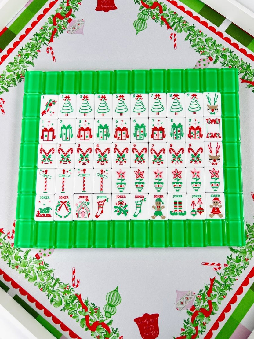 Holiday Mahjong tiles arranged in rows on green racks over the Holly Jolly Christmas mat, with Christmas tree, gift, candy cane, ornament, and holiday character designs.