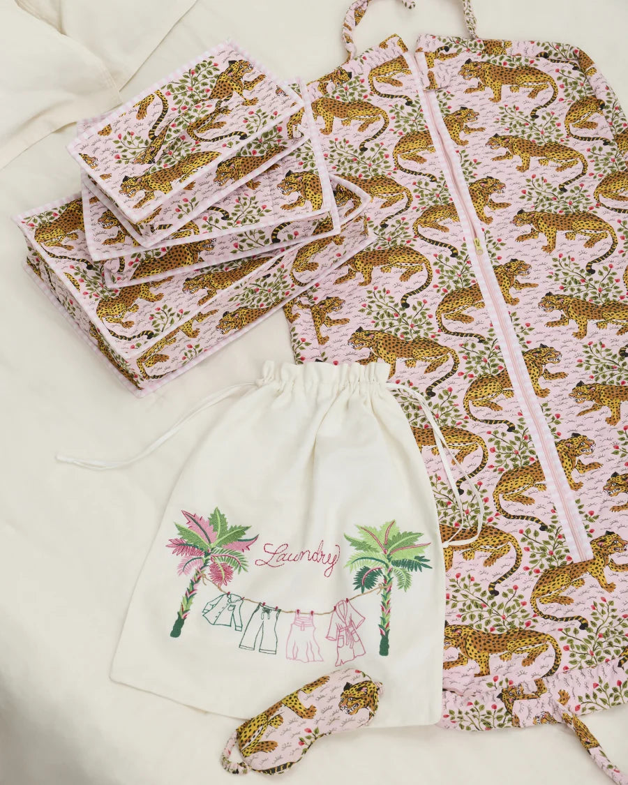 Pink leopard-print packing cubes and garment bag arranged beside an embroidered laundry bag and matching eye mask.